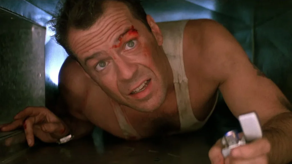 Bruce Willis rasteja por um duto de ventilação, com lanterna na mão e olhar atento, em cena de Duro de Matar.
