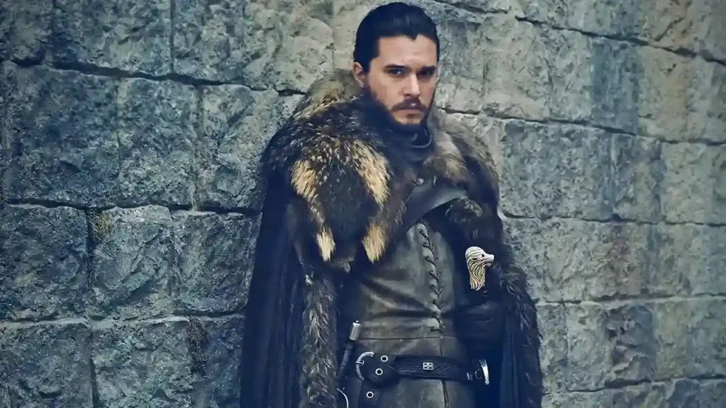 Kit Harington em traje de couro e capa de pele, encostado em uma muralha de pedra, em cena de Game of Thrones.