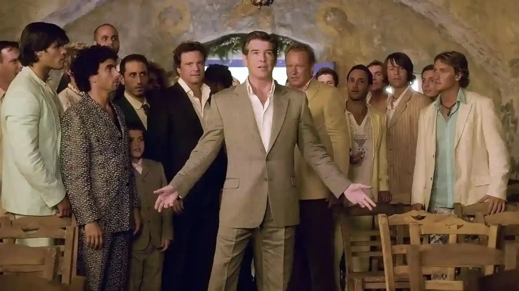 Pierce Brosnan de braços abertos em cena de Mamma Mia!, cercado por elenco masculino em taverna iluminada.