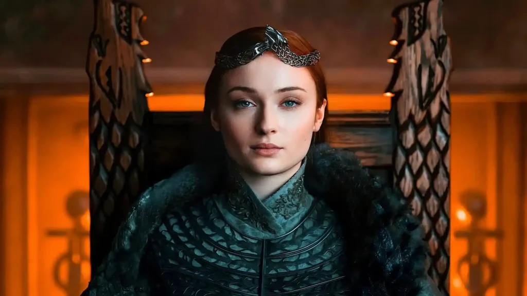 Sophie Turner sentada em trono de madeira, usando coroa prateada e armadura escura.