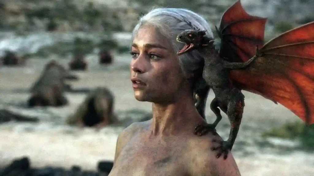 Emilia Clarke caracterizada como Daenerys Targaryen, com cabelo preso e expressão séria diante do trono.
