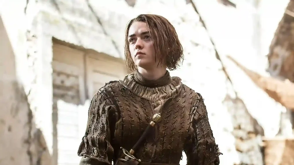 Maisie Williams em trajes de batalha, com espada à cintura, em ambiente externo de pedra