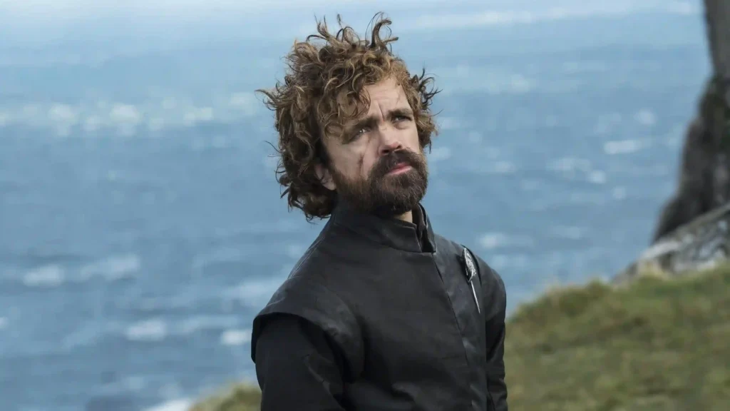 Peter Dinklage com expressão séria e olhar distante, em cenário costeiro de Game of Thrones.