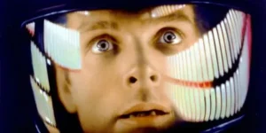 Close-up do rosto de um astronauta com olhos arregalados, refletindo luzes coloridas em seu capacete, em uma cena icônica do filme 2001: Uma Odisseia no Espaço.