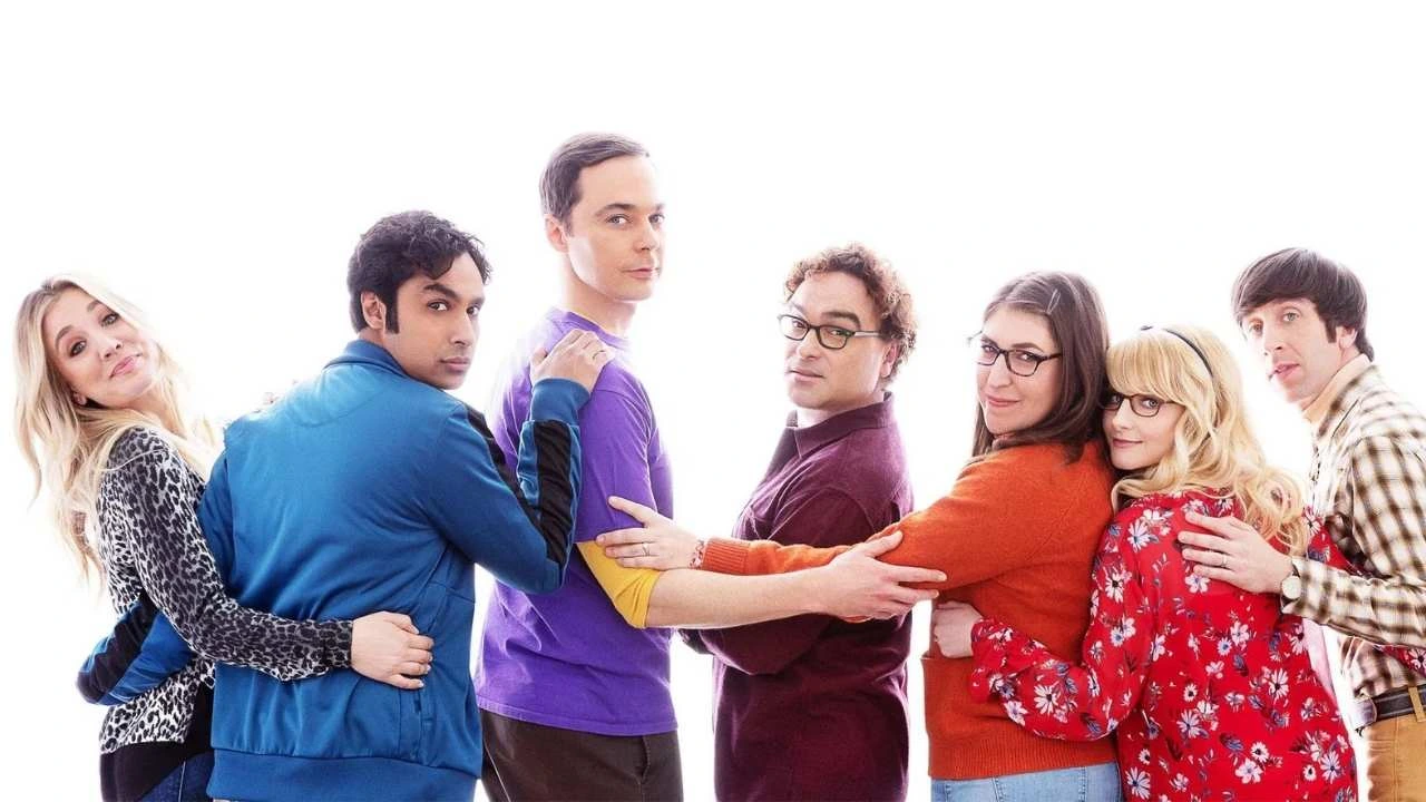 Elenco completo de The Big Bang Theory em fila, abraçados sobre fundo branco. Penny (blusa de oncinha), Raj (casaco azul), Sheldon (camiseta roxa), Leonard (suéter bordô), Amy (suéter laranja), Bernadette (vestido vermelho) e Howard (camisa xadrez).