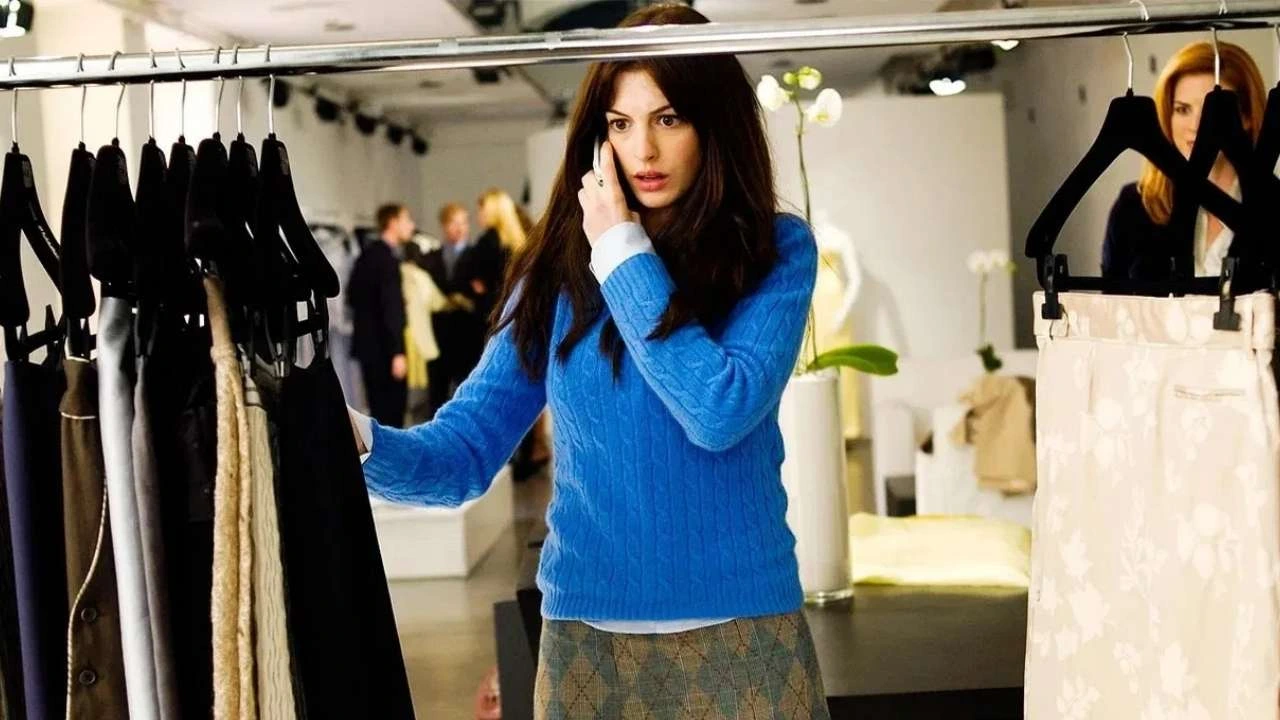 Anne Hathaway como Andy Sachs, vestindo um suéter azul de tricô e saia xadrez, falando ao telefone em uma loja de roupas elegante. Ao redor dela, cabides com roupas pretas e brancas penduradas em araras, enquanto pessoas conversam ao fundo em um ambiente de moda.