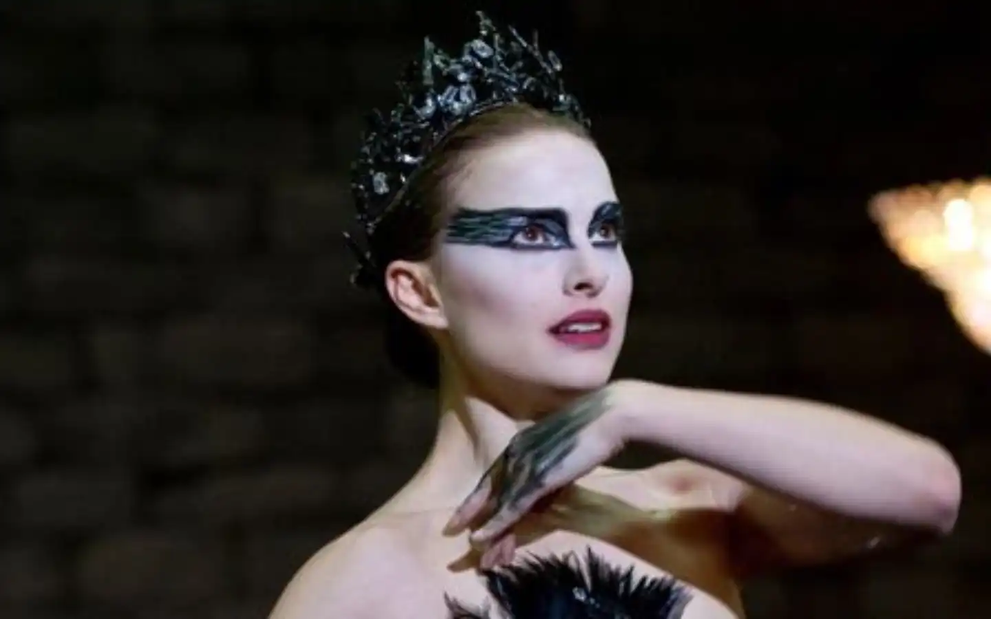 Imagem do filme Cisne Negro - Mulher com maquiagem dramática preta ao redor dos olhos e coroa escura, usando figurino de balé preto com penas, em uma performance intensa e expressionista.