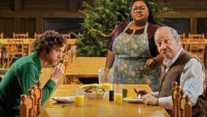 Cena do filme Os Rejeitados com três personagens em um refeitório: um jovem sentado comendo, uma cozinheira com avental e expressão séria ao fundo, e um senhor idoso sentado à mesa.