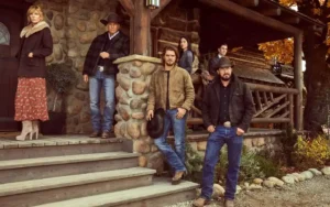 Elenco da série 'Yellowstone' reunido em frente a uma casa de madeira com colunas de pedra. Personagens vestem roupas típicas de fazenda, incluindo chapéus e botas.