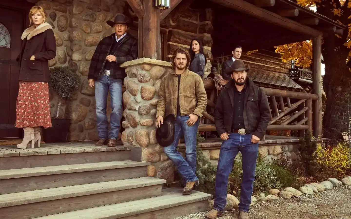 Elenco da série 'Yellowstone' reunido em frente a uma casa de madeira com colunas de pedra. Personagens vestem roupas típicas de fazenda, incluindo chapéus e botas.