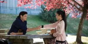 Una pareja de adolescentes se sienta en lados opuestos de una mesa de piedra bajo un árbol en flor, intercambiando un apretón de manos, en una escena romántica de la película 'A todos los chicos de los que me enamoré