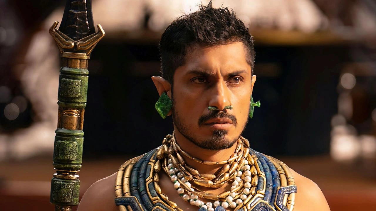 Primeiro plano de Tenoch Huerta como Namor em Pantera Negra: Wakanda Para Sempre, com expressão séria e determinada. Ele usa adornos de orelha verdes, colares de contas brancas e detalhes azuis típicos da cultura maia, representando o poderoso governante do reino submarino de Talokan.