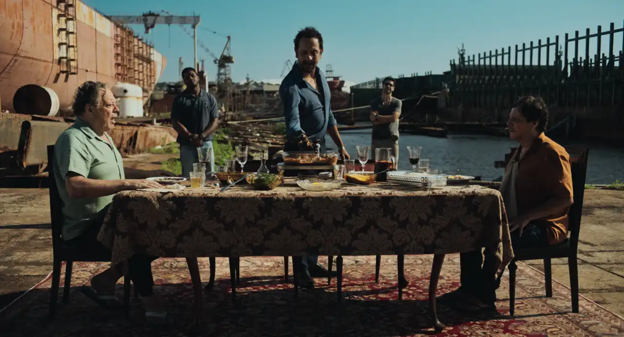 Irandhir Santos serve comida em uma mesa posta ao ar livre, cercado por personagens, em cena de Os Enforcados