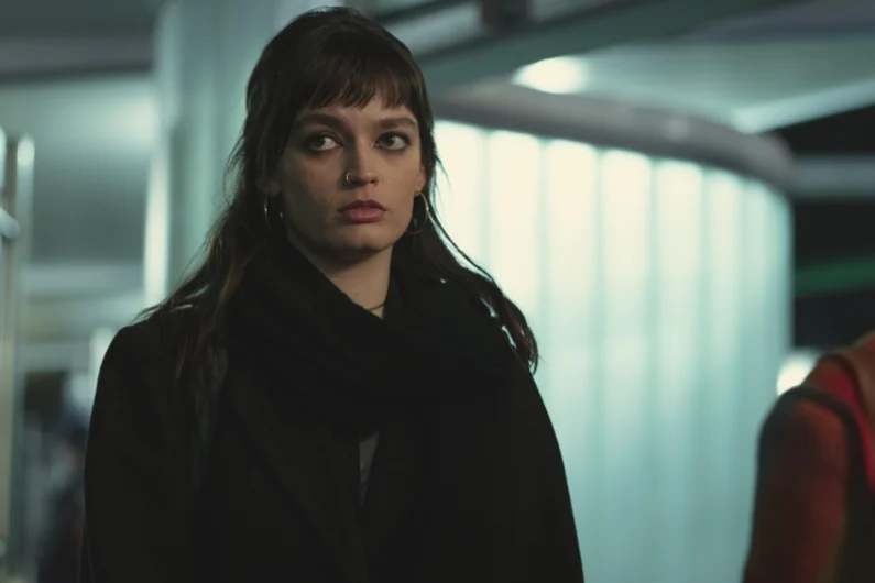 Emma Mackey with a pensive expression, wearing a dark coat, in an urban environment.