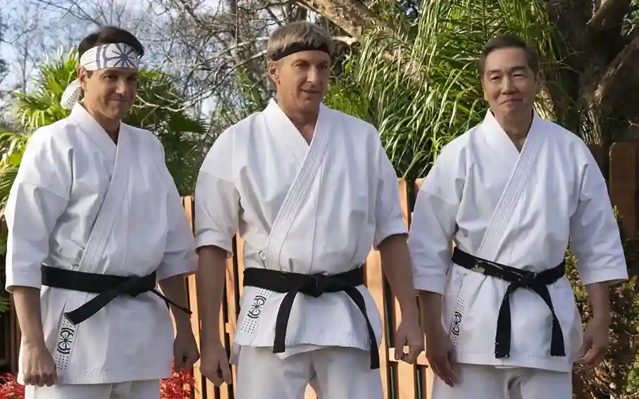 Três senseis de Cobra Kai usando uniformes brancos de karatê com faixas pretas, em pé lado a lado em um jardim. À esquerda, Johnny Lawrence com bandana branca, no centro seu antigo sensei John Kreese com bandana preta, e à direita o Sr. Miyagi, todos em postura formal diante de vegetação tropical.(Créditos: TMDB)