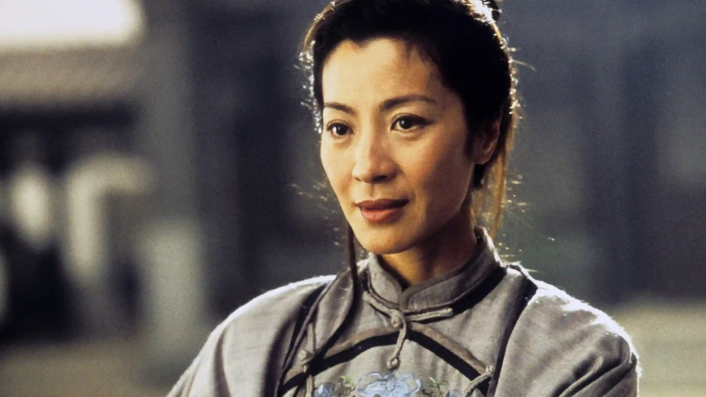 Primer plano de Michelle Yeoh como Yu Shu Lien en El tigre y el dragón (2000), con expresión serena, cabello recogido y vistiendo un traje tradicional chino gris con cuello mandarín, iluminada suavemente contra un fondo desenfocado. (Credito: TMDB)