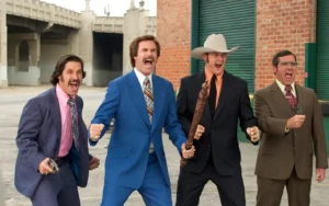 Four men in colorful 1970s suits, standing in a city alleyway, each holding improvised weapons and striking dramatic poses. Promotional scene from the comedy film 'Anchorman: The Legend of Ron Burgundy'. (Credits: TMDB)