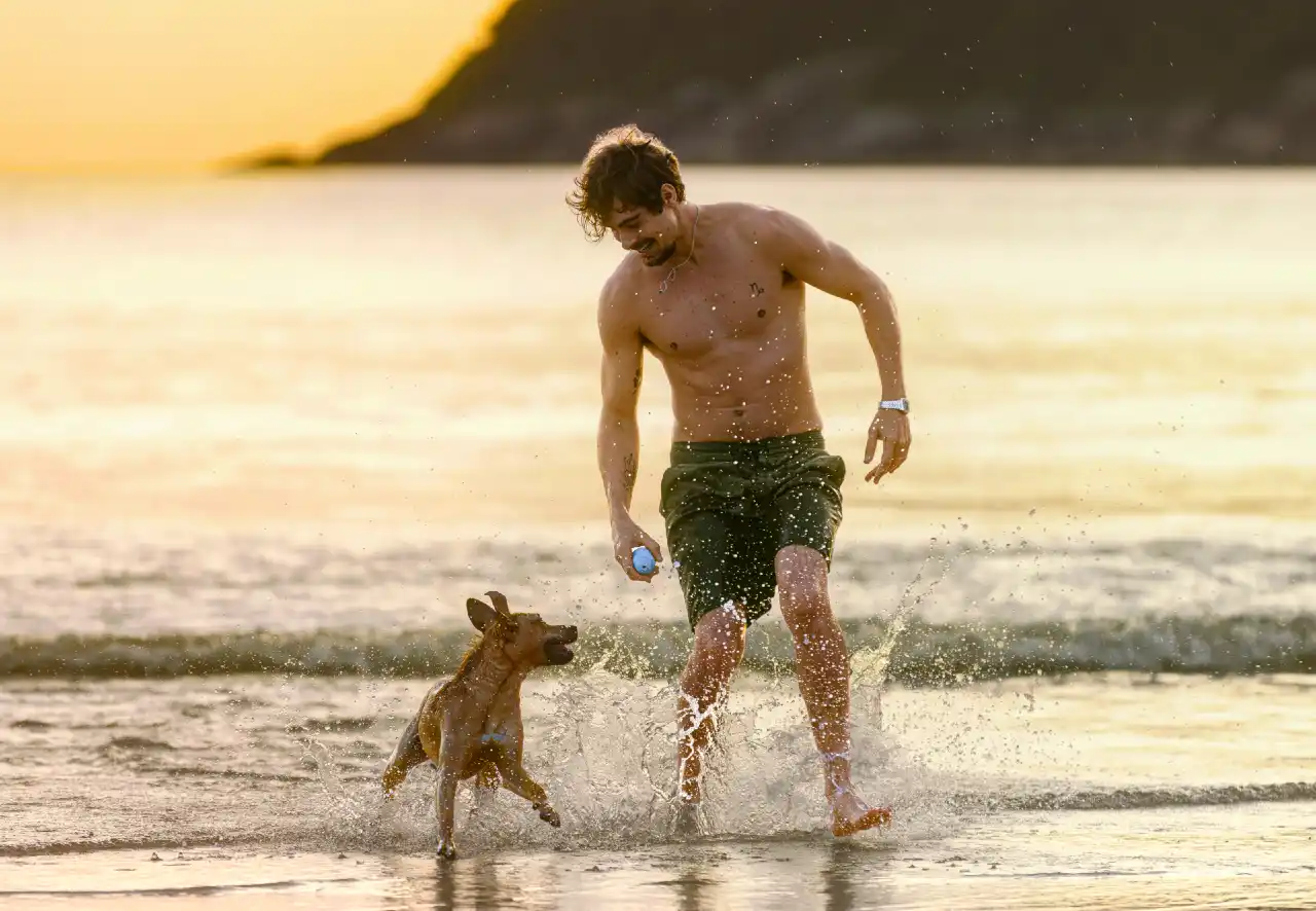 O ator Rafael Vitti, sem camisa e de bermuda verde, brinca com o cachorro vira-lata Caramelo na beira do mar, em cena de pôr do sol do filme Caramelo