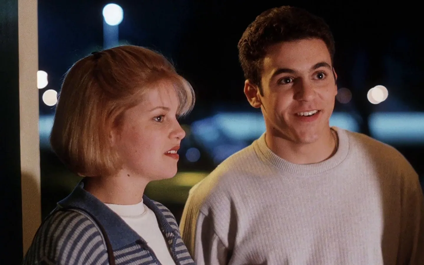 Two teenagers standing side by side at the entrance of a house at night, illuminated by soft light from inside; the girl wears a striped sweater and the boy a light-colored sweatshirt. Scene from the film 'No One Would Tell'. (Créditos: TMDB)
