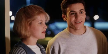 Two teenagers standing side by side at the entrance of a house at night, illuminated by soft light from inside; the girl wears a striped sweater and the boy a light-colored sweatshirt. Scene from the film 'No One Would Tell'. (Créditos: TMDB)
