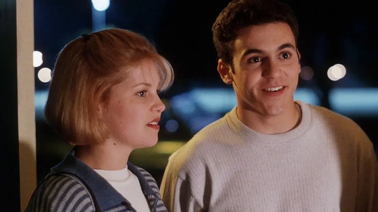 Two teenagers standing side by side at the entrance of a house at night, illuminated by soft light from inside; the girl wears a striped sweater and the boy a light-colored sweatshirt. Scene from the film 'No One Would Tell'. (Créditos: TMDB)